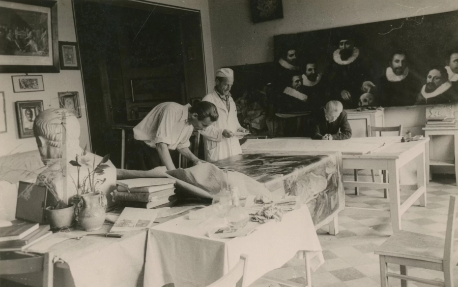 Melnbalts fotoattēls, kurā redzamas vairākas personas, kas iesaistītas dažādās aktivitātēs telpā, kas piepildīta ar mākslas darbiem un klasisku dekoru. Viens cilvēks uz galda stiepj lielu auduma gabalu, bet cits raksta vai zīmē pie atsevišķa galda. Telpu 