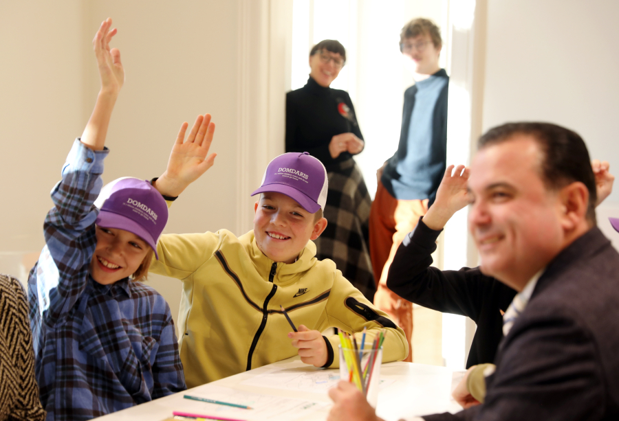 Skolēni spilgti apgaismotā klasē ar paceltām rokām, sēž pie galdiem ar papīriem un zīmuļiem, piedalās klases aktivitātē vai diskusijā.