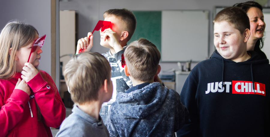 Attēlā redzami pieci 5. klases skolēni - meitenes un zēni. Divi no viņiem pie acīm ir pielikuši lielas sarkanas brilles. Vienam zēnam uz džempera ir uzraksts 