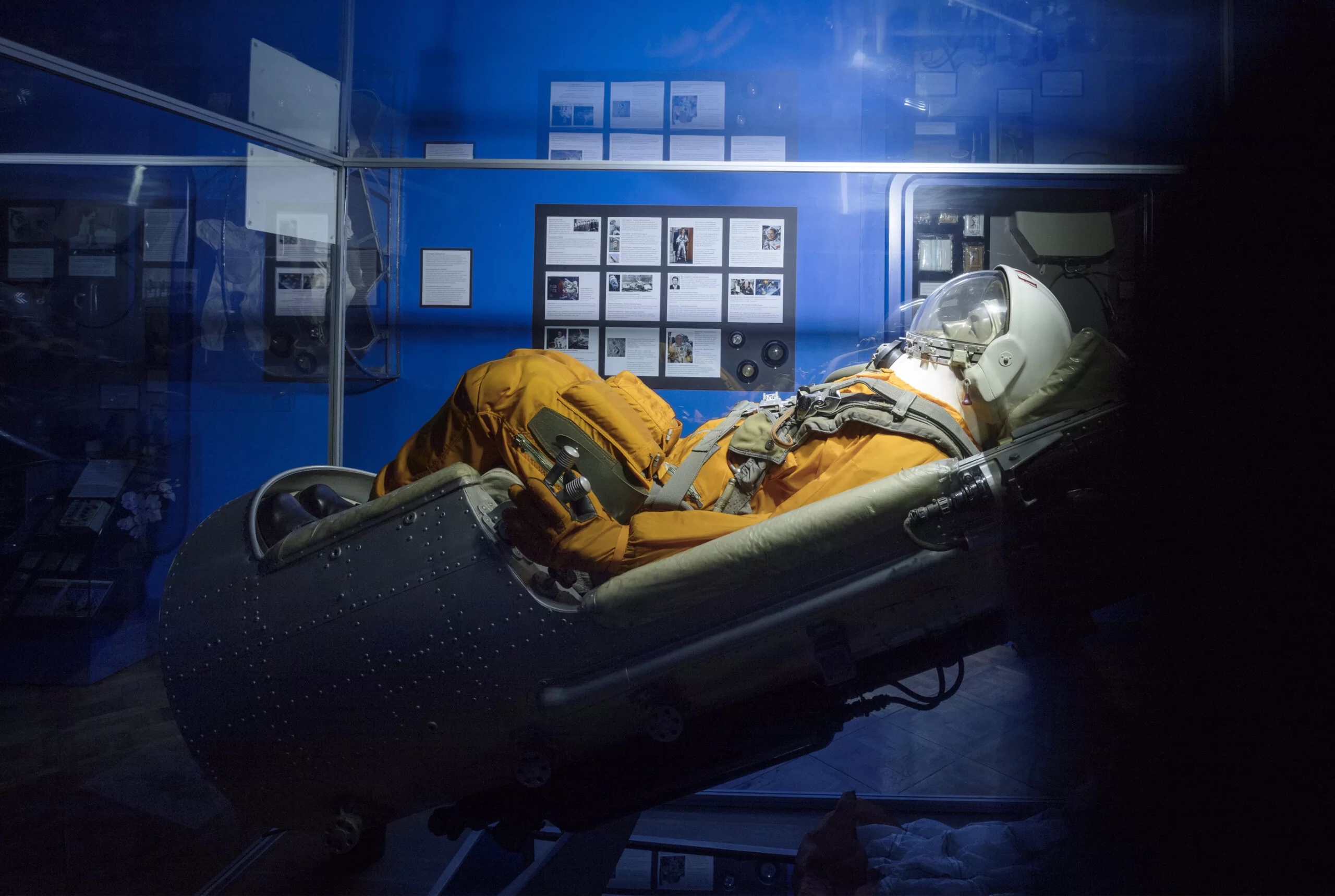 Fotogrāfija no muzeja kosmosa ekspozīcijas, kurā redzams astronauta tērpā ģērbts manekens uz krēsla. Fonā zilas sienas uz kurām atrodas dažādas informatīvas planšetes.