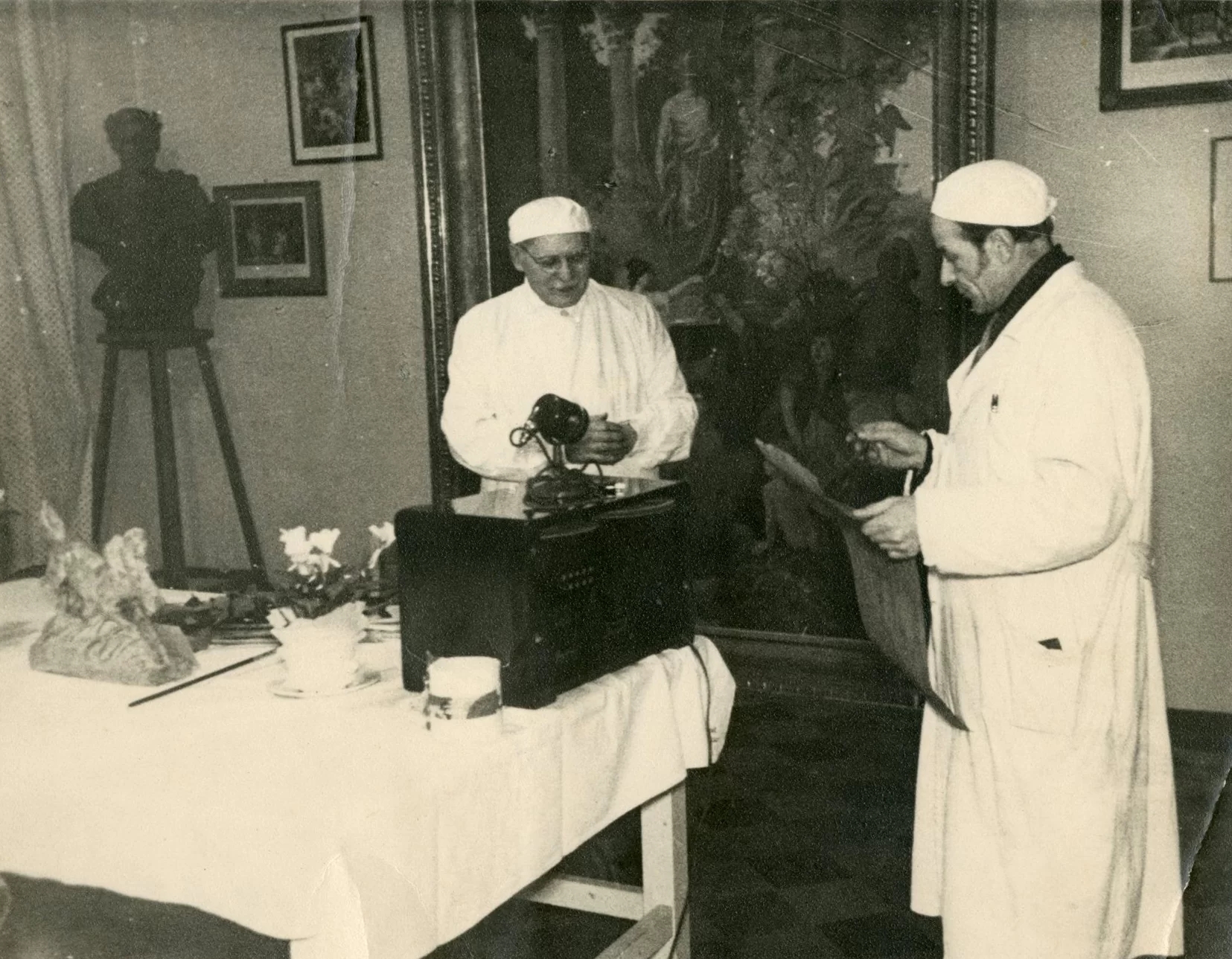 Fotogrāfija no ekspozīcijas uzstādīšanas, kurā redzami divi cilvēki blakus balss ierakstīšanas ierīcei. Uz galda redzami daži eksponāti, kā arī uz sienām atrodas vairākas gleznas.