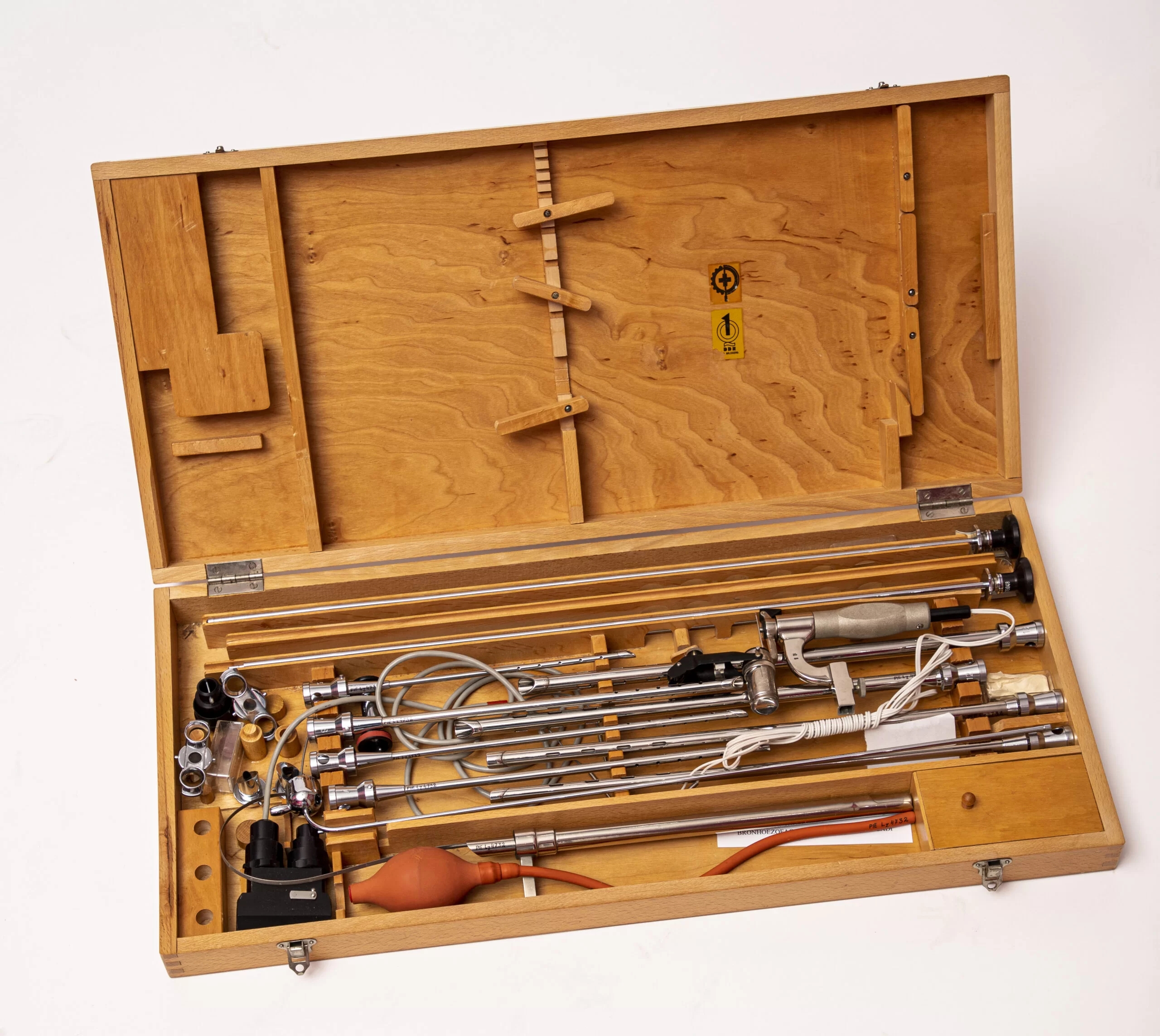 Bronhoskopijas instrumentu komplekta attēls. Instrumenti atrodas gaišā koka kastē un katram instrumentam redzams speciāli pielāgotas atbalsta detaļas.